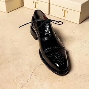 May include: A black patent leather oxford shoe featuring a cap toe with decorative brogue perforations, a lace-up closure with thin black laces, and a stacked heel. The interior lining is dark brown, and the letter T is visible inside.