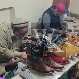 May include: Two artisans working on footwear. A pair of brown cowboy boots with musical notes and guitar designs are in the foreground. Other shoes in various colors, including red, tan, and green, are on a table. Tools and materials are visible.