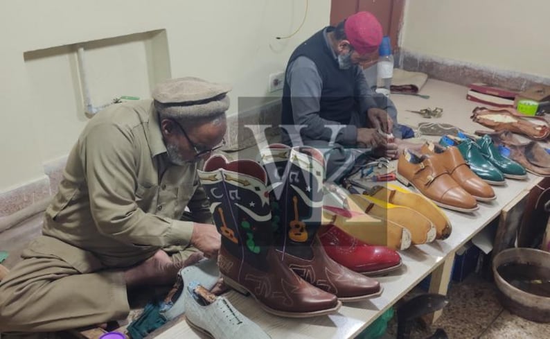 May include: Two artisans working on footwear. A pair of brown cowboy boots with musical notes and guitar designs are in the foreground. Other shoes in various colors, including red, tan, and green, are on a table. Tools and materials are visible.