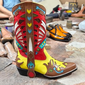 Puede incluir: Una bota vaquera de cuero multicolor con una caña roja vibrante que presenta recortes geométricos en blanco, amarillo y azul. La parte inferior es de color amarillo brillante con superposiciones de cuero marrón, un trébol verde y un ave azul.