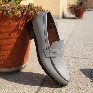 May include: A gray leather loafer with a black sole and heel. The shoe features a strap across the vamp and white stitching around the edges. The shoe is leaning against a terracotta pot.