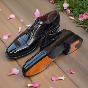 May include: A pair of polished black leather dress shoes with a cap toe design. The shoes have a glossy finish, intricate stitching, and a wooden sole. The image shows the shoes from multiple angles, with pink flower petals scattered around them.