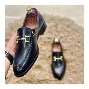 K&ouml;nnte beinhalten: Schwarze Leder-Loafer mit goldfarbener Schnalle. Die Schuhe haben ein klassisches Design mit runder Spitze und niedrigem Absatz. Das Bild zeigt zwei Loafer, einer im Fokus und einer im Hintergrund.