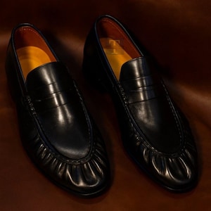 Peut inclure: Une paire de mocassins en cuir noir au design classique. Les chaussures pr&eacute;sentent un bout arrondi, une bride penny et une semelle int&eacute;rieure jaune. Les mocassins sont pr&eacute;sent&eacute;s sur une surface marron.