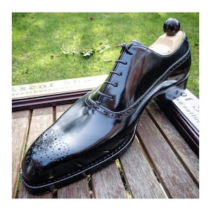 May include: A polished black leather oxford shoe featuring a cap toe with decorative brogue perforations, a closed lacing system with thin black laces, and a wooden shoe tree inserted inside to maintain the shape of the footwear.