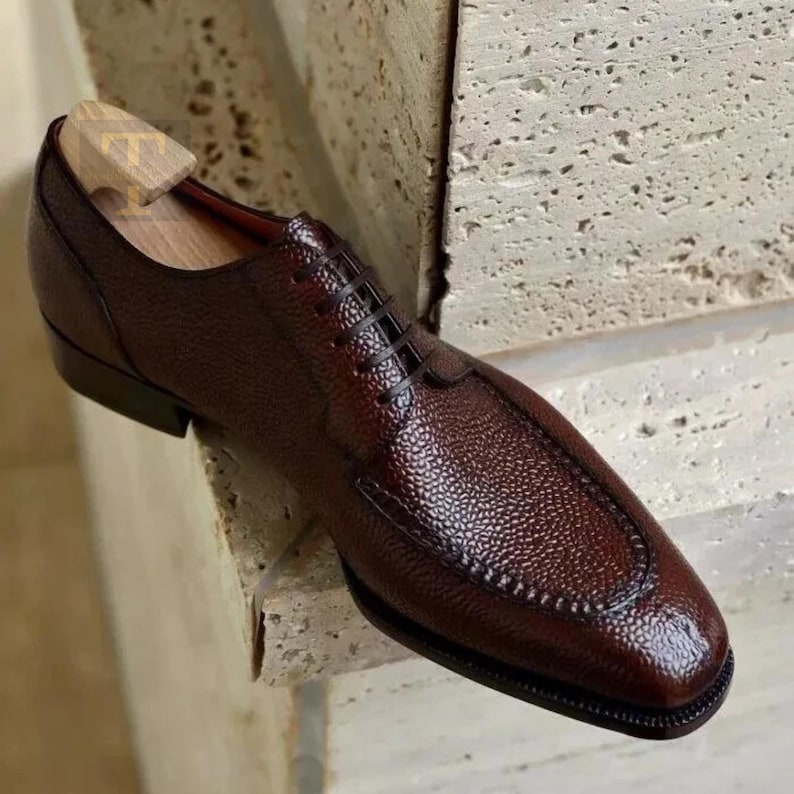 May include: A brown leather dress shoe with a textured surface and a classic lace-up design. The shoe features a rounded toe and a wooden shoe tree inside. The shoe is a rich, dark brown color.
