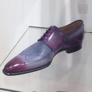 May include: A single, two-tone leather dress shoe. The shoe transitions from a deep burgundy at the toe to a light blue-grey at the midfoot. The shoe has a wingtip design with burgundy laces and a black sole.