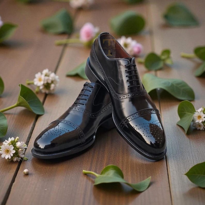 May include: A pair of polished black leather dress shoes with intricate detailing, including perforations and a lace-up design. The shoes are set on a wooden surface, with scattered greenery and small white flowers.