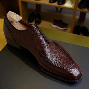 May include: A single, brown leather dress shoe with a textured surface and a wooden shoe tree inserted. The shoe has a classic lace-up design and a stitched sole. The background shows a shelf with other shoes.