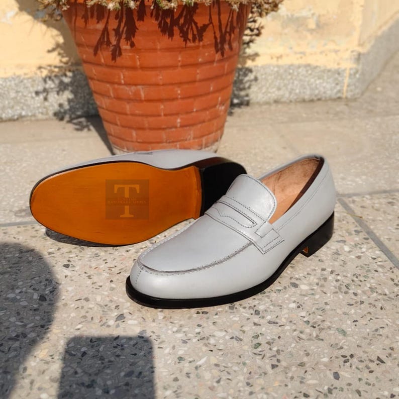 May include: A pair of light gray leather loafers with a classic design. The shoes have a rounded toe, a strap across the vamp, and a black sole. The sole of one shoe is visible, showing a brown leather sole.
