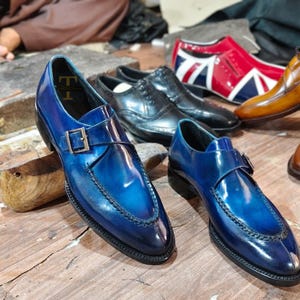 May include: A pair of blue leather dress shoes with a buckle fastening. The shoes have a glossy finish and a stitched design around the edges. Other shoes in various colours are visible in the background.