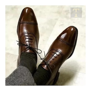 Op de afbeelding: Een paar bruine leren schoenen met een glanzende afwerking. De schoenen hebben een klassiek Oxford-ontwerp met decoratieve perforaties en veters. De schoenen worden gedragen met grijze sokken.
