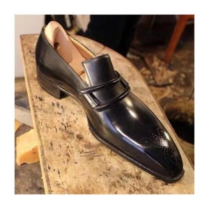 May include: A black leather loafer with a glossy finish. The shoe features a decorative strap across the vamp and a perforated design on the toe. The shoe is placed on a wooden surface.