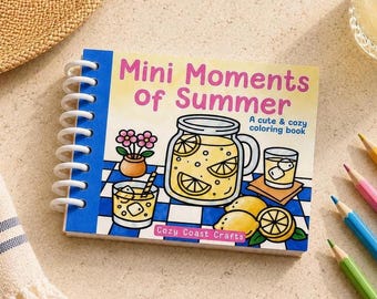 May include: A spiral-bound coloring book titled "Mini Moments of Summer" with a blue spine. The cover features illustrations of lemonade, lemons, and flowers. The text "A cute & cozy coloring book" is also on the cover. Colored pencils are scattered around the book.