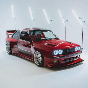 May include: A modified deep red BMW E30 coupe featuring a wide body kit, a large rear wing, and custom multi-spoke chrome wheels. The front end displays bright circular LED headlights, a black grille, and a front splitter. Text reads THE LOWDOWN.
