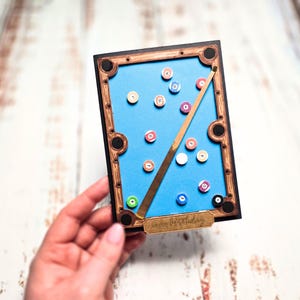 May include: A rectangular card featuring a miniature pool table design. The card has a blue playing surface with numbered pool balls, a wooden frame, and a cue stick. The card has the words "Happy Birthday" on it.
