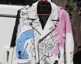 Könnte beinhalten: Weiße Lederjacke mit schwarzen und rosa Akzenten, handbemalt mit Kunstwerken und dem Wort "DIE" auf der linken Schulter. Die Jacke hat einen Reißverschluss und einen Gürtel. Der rechte Ärmel hat einen rosa Farbverlauf.