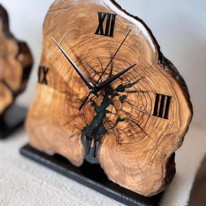 May include: A unique wooden clock with a natural edge, featuring black Roman numerals and hands. The clock face is made from a cross-section of a tree trunk, with a dark blue resin filling a natural void. It sits on a black base.