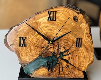 May include: A unique wooden clock with a natural edge. The clock face is made from a cross-section of a tree trunk, featuring visible growth rings and a teal-colored resin inlay. Black Roman numerals mark the hours, and black hands indicate the time. The clock sits on a black base.