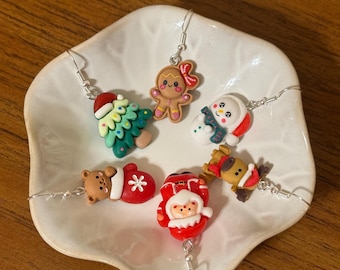 May include: A collection of Christmas-themed earrings displayed on a white plate. The earrings feature festive designs like a Christmas tree, gingerbread person, snowman, bear in a mitten, Santa Claus, and a reindeer. Each earring has a silver hook.