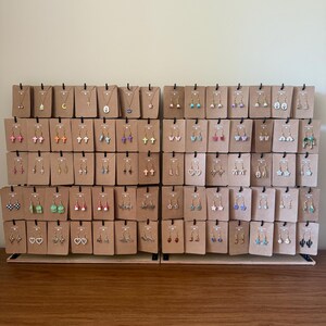 May include: A display of various earrings and necklaces on brown cardboard backing. The jewelry includes cross, heart, star, and butterfly designs in gold, silver, and colorful accents. The earrings are displayed on a wooden surface.