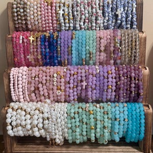 May include: A wooden display stand holds multiple beaded bracelets in various colors, including pink, blue, white, and green. Some bracelets feature cross charms. The beads vary in size and material, creating a diverse collection of jewelry.