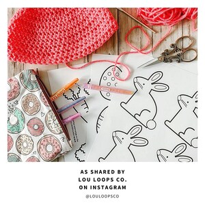 May include: A set of printable coloring pages featuring bunny rabbits, an egg, and a tree. The pages are on white paper and are ready to be colored. The image also includes a pink crocheted hat, a pencil case with a donut pattern, and a pair of scissors.