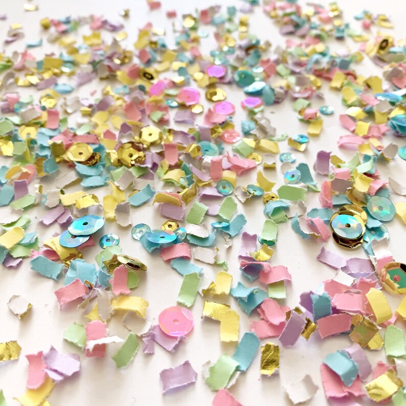 May include: A scattering of colorful confetti and sequins in pastel shades of pink, blue, yellow, and green. Gold metallic accents add a touch of sparkle. The confetti is in small, irregular shapes, creating a festive and celebratory look.