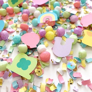 May include: A colorful assortment of confetti, paper cutouts, and small, round, textured objects. The confetti includes gold, pink, and green pieces. Paper cutouts include a rainbow, a horseshoe, and a four-leaf clover. The round objects are in pastel colors.