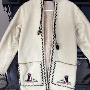Peut inclure: Un manteau à capuche couleur crème avec une bordure en fausse fourrure, des détails décoratifs en point de surjet noir le long de l'ouverture et des poches, et des motifs brodés de macareux sur les deux poches avant avec des perles en bois.