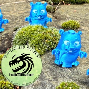 Può includere: Diverse statuette blu stampate in 3D di una creatura simile a un cartone animato con grandi occhi e braccia piccole sono disposte su una superficie rocciosa con muschio. Le statuette hanno un aspetto strutturato. È visibile anche un logo circolare con la scritta "AUTHORIZED SELLER" e "hollowmaker".