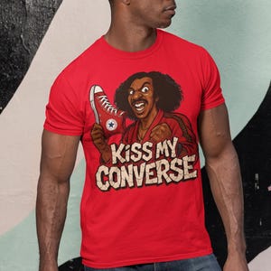 May include: Red t-shirt with a graphic of a smiling man holding a red Converse shoe. The text "Kiss My Converse" is printed below the graphic in a distressed font. The man is wearing sunglasses and blue jeans.