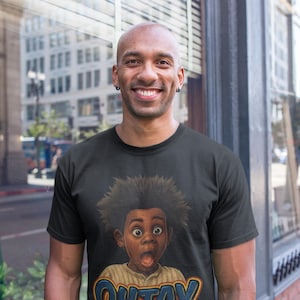 Puede incluir: Camiseta negra con una ilustración de dibujos animados de un niño sorprendido con ojos grandes y una expresión de asombro. El texto "OHTAY" se muestra en azul y naranja. La persona que lleva la camiseta está sonriendo.