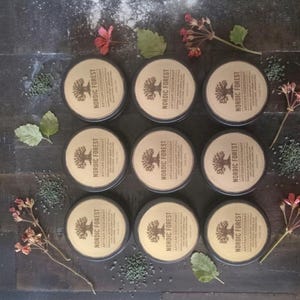May include: Nine circular metal tins of artisan deodorant are arranged on a dark wooden surface. Each tin features a tan label with the words "NORDIC FOREST" and a tree graphic. Sprigs of red flowers and green leaves are scattered around the tins.