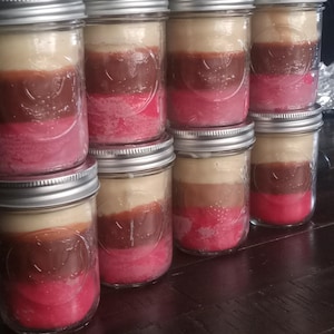 Puede incluir: Una colección de ocho velas en capas en frascos de vidrio con tapas plateadas. Cada vela tiene una combinación de colores rosa, marrón y beige. Las velas están apiladas sobre una superficie de madera oscura.