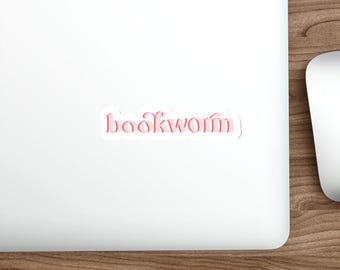 Peut inclure: Un autocollant blanc avec le mot "bookworm" en rose, en écriture cursive. L'autocollant est sur un ordinateur portable blanc, avec un bureau en bois et un stylet blanc en arrière-plan. L'autocollant a une bordure blanche.
