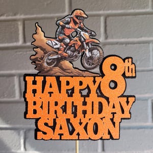 May include: Orange and black cake topper with a motocross rider on a dirt bike. The text reads "Happy Birthday Saxon 8th". The topper has glitter accents and is mounted on a wooden stick, perfect for an eighth birthday.