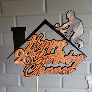 May include: A black and orange glitter cake topper featuring a house roof silhouette with a chimney and a construction worker figure. The text reads Happy 26th Birthday Chance in a layered script font, supported by a single thin wooden stick.