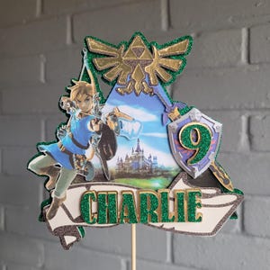 May include: A decorative cake topper featuring a layered design with a green glittery Triforce symbol at the top, a central image of a character with a bow, a castle, a shield with the number 9, and a banner with the name CHARLIE in green glitter.