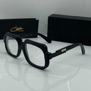Peut inclure: Lunettes Cazal Legends noires avec une monture carrée épaisse. Les lunettes ont des verres transparents et un logo Cazal doré sur la branche. Un étui et une boîte noirs avec le logo Cazal sont également visibles.