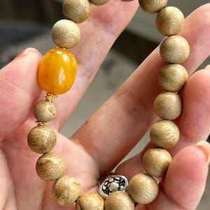 Op de afbeelding: Een armband bestaande uit ronde, lichtbruine houten kralen met zichtbare houtnerven. De armband bevat één gladde, ovaalvormige gele barnstenen kraal, geaccentueerd door een kleine goudkleurige tussenkraal en een zilverkleurige bedel met reliëf.