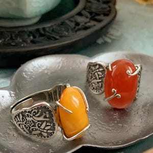 May include: Three silver rings with ornate designs and gemstone settings. One ring features an oval, amber-colored stone, while another has a vibrant red stone. The rings rest on a decorative silver tray, showcasing the jewelry's intricate details.
