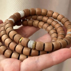 May include: A wooden bead bracelet in natural brown. The bracelet has cylindrical beads strung together, with white and gold accents. The beads are approximately 1.3 cm in diameter. The bracelet is wrapped around a hand.
