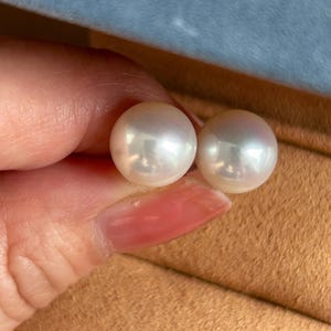 May include: A pair of round, white stud earrings featuring smooth, lustrous pearls with subtle iridescent highlights. The pearls exhibit a high-gloss finish and are spherical in shape, reflecting light across their polished surfaces.