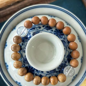 May include: A wooden bead bracelet with clear, round accent beads. The bracelet is displayed on a white and blue patterned ceramic plate, with a small white teacup in the center. The beads are a light brown color.
