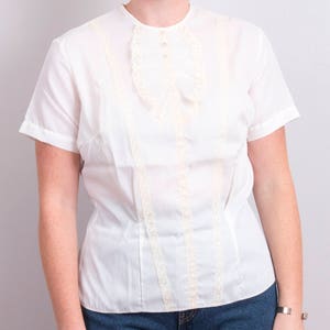 Könnte beinhalten: Eine weiße Bluse mit kurzen Ärmeln, rundem Ausschnitt und dekorativer Spitzenborte. Die Bluse hat einen gerüschten Akzent auf der Brust und vertikale Spitzendetails auf der Vorderseite. Der Stoff scheint leicht zu sein.