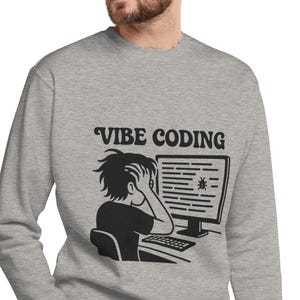 Puede incluir: Sudadera gris con el texto "VIBE CODING" sobre un gráfico negro de una persona frente a una computadora. La persona se agarra la cabeza con frustración, con código en la pantalla.