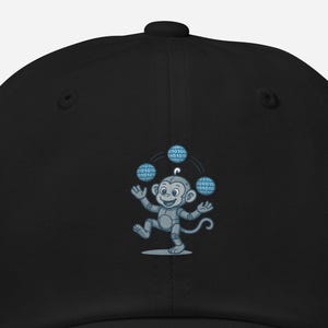 May include: Black baseball cap with a cartoon monkey juggling three blue and white globe-shaped balls. The monkey is light blue and grey, with a smiling expression. The cap has a curved peak and a button on top.
