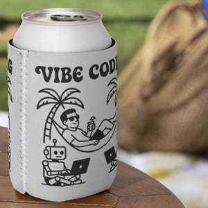 Op de afbeelding: Een witte blikjeskoeler met de tekst "Vibe Code". Het ontwerp toont een persoon die ontspant in een hangmat onder een palmboom, een robot en een laptop. De koeler staat op een houten tafel.