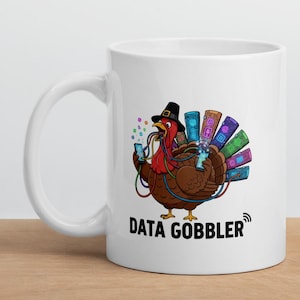 Könnte beinhalten: Weiße Keramik-Tasse mit einem Cartoon-Truthahn, der einen Hut trägt und ein Smartphone hält. Der Truthahn hat bunte Daten-Schwanzfedern und der Text "DATA GOBBLER" ist auf der Tasse aufgedruckt.